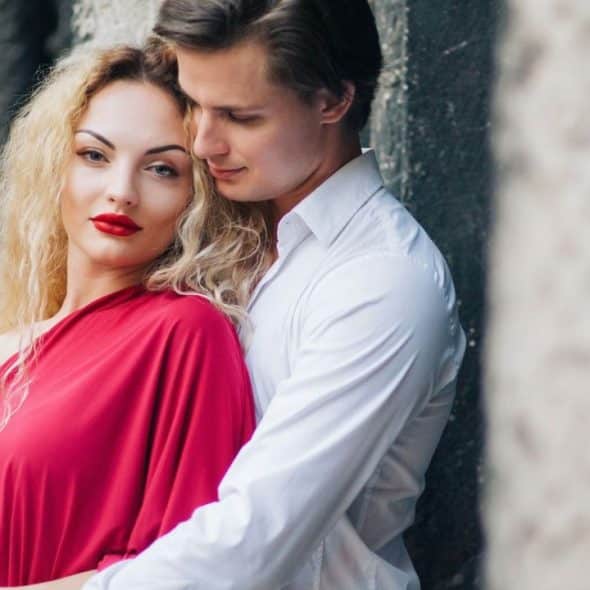 A man in a white shirt embraces a blonde woman wearing a bright red dress.