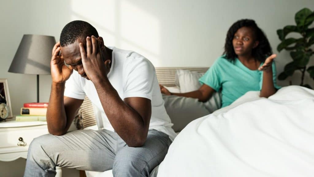 A man sits on a bed holding his head while a woman gestures behind him.