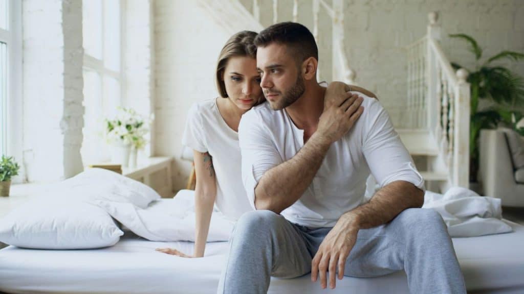 A woman rests her hand on the shoulder of a man sitting on a bed.