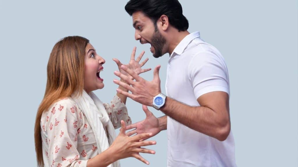 A man sits looking away while a woman behind him gestures with her hands.