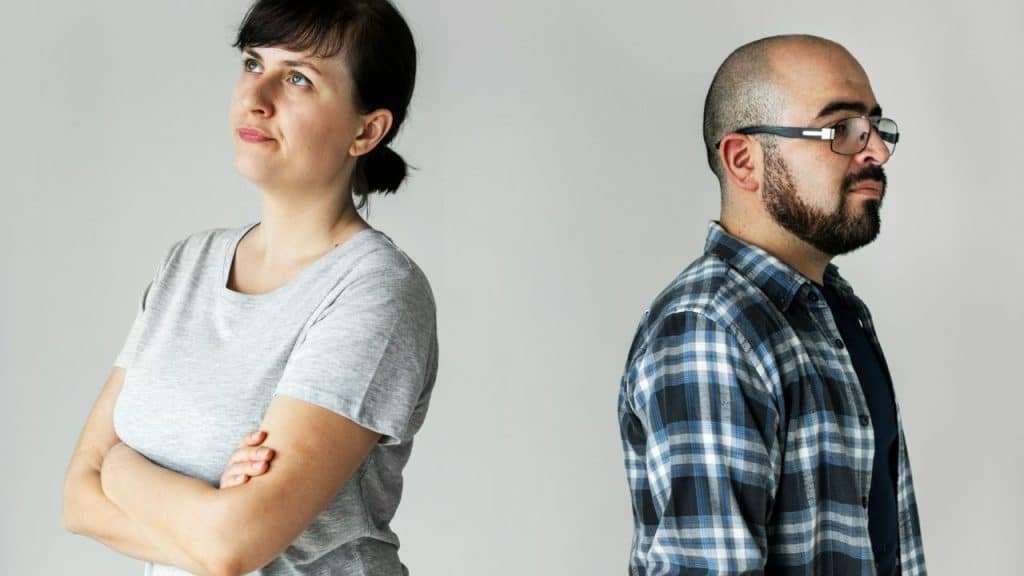 A woman with crossed arms and a man in glasses stand back to back.