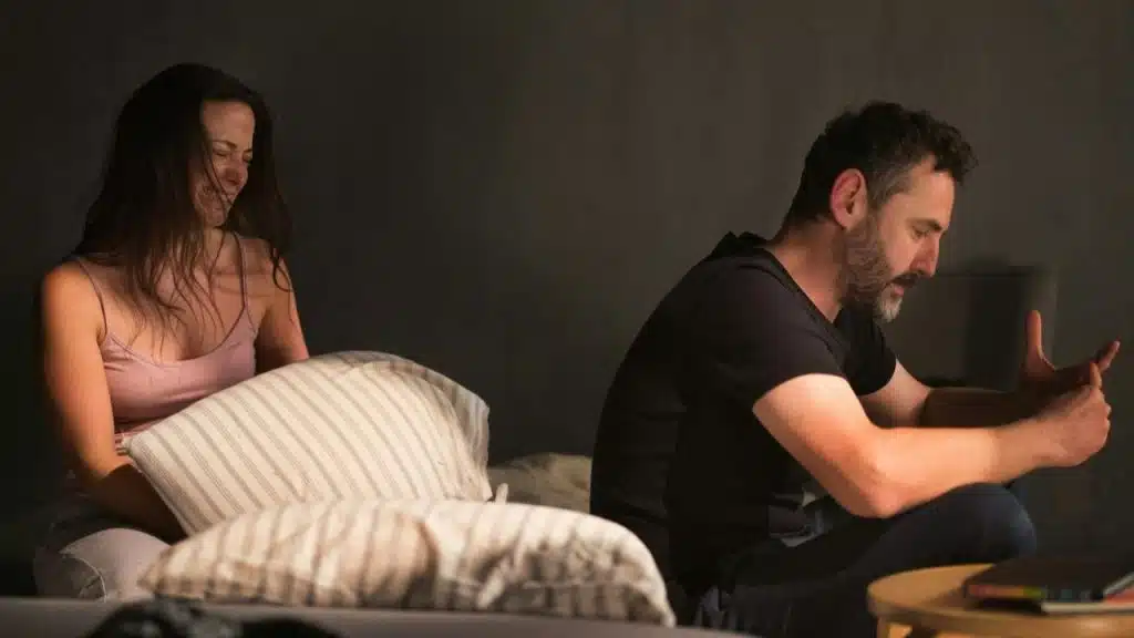 A woman sits on a bed looking upset while a man sits nearby looking down.