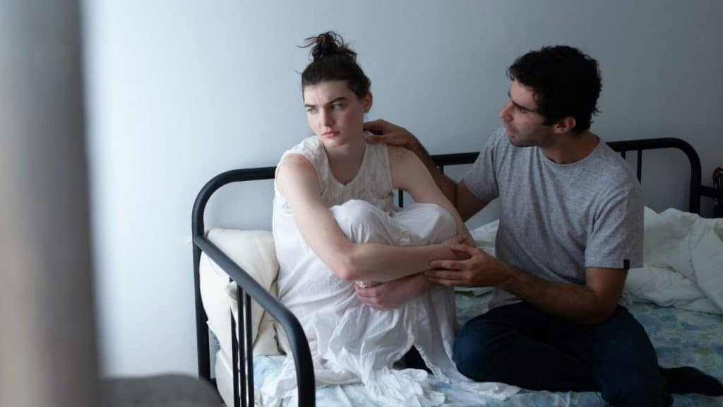 A woman sits on a bed looking away while a man rests a hand on her.