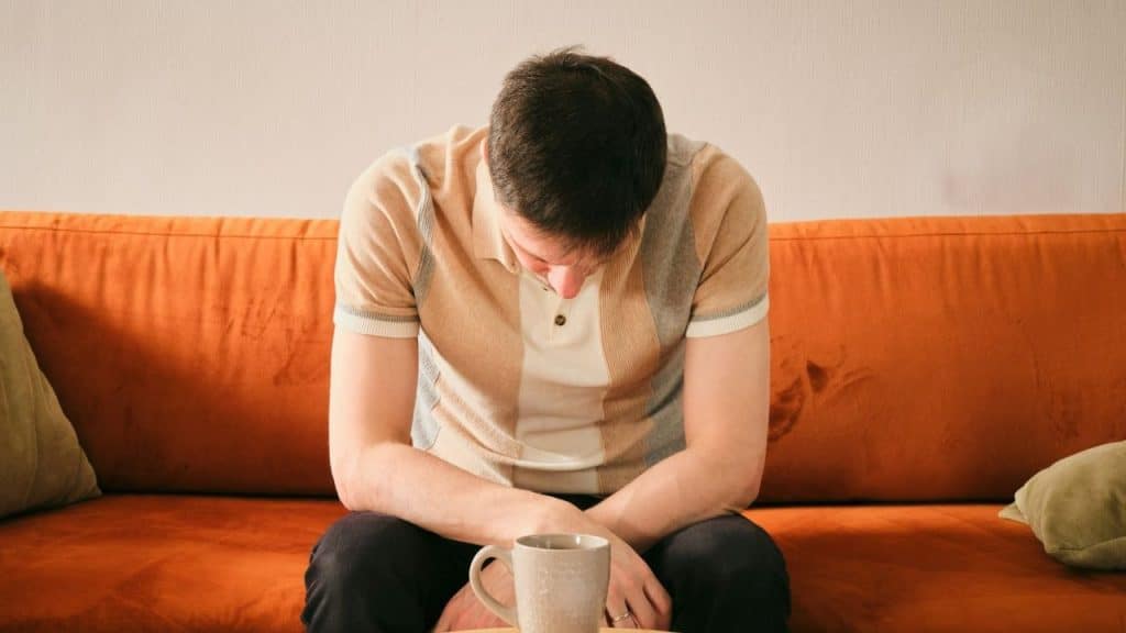 A man sits on an orange sofa with his head bowed over a light-colored mug.