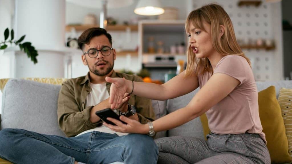 A woman gestures toward a smartphone held by a man sitting on a sofa.