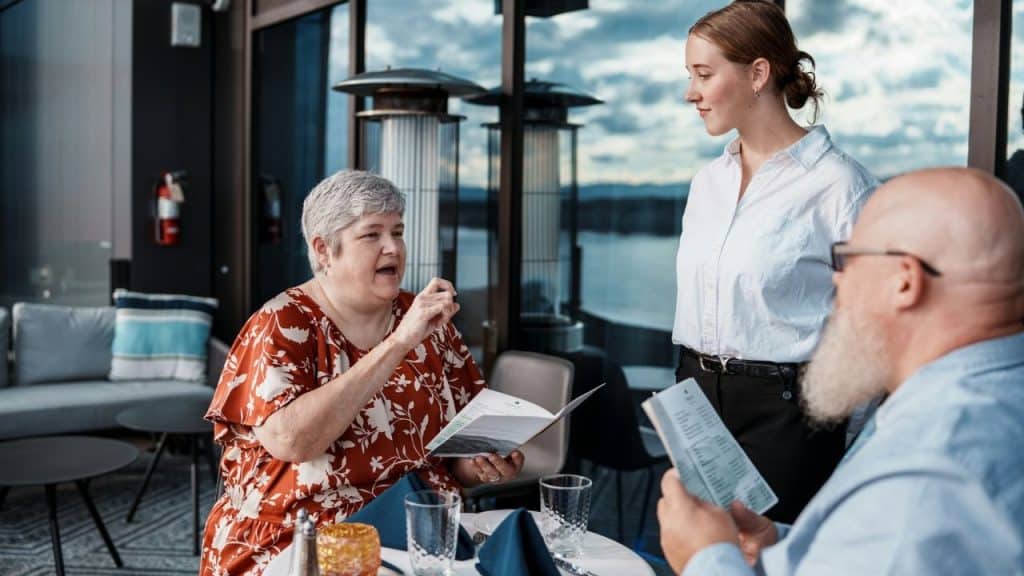 A woman in a floral dress speaks to a server standing by a restaurant table.