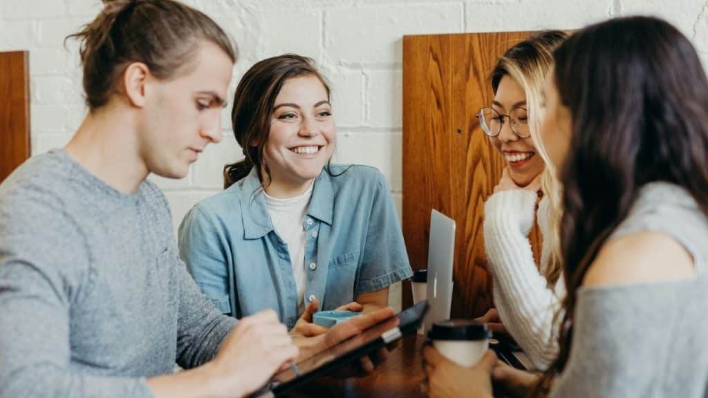 Four people sit at a table with coffee and a tablet, smiling and talking together.