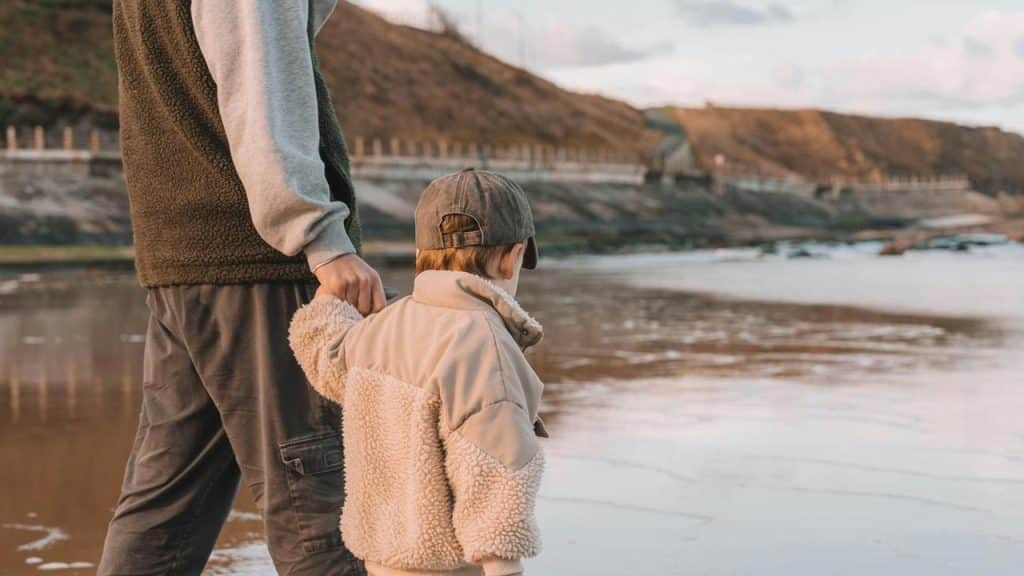 A child holding an adultโs hand while walking along a beach at low tide.