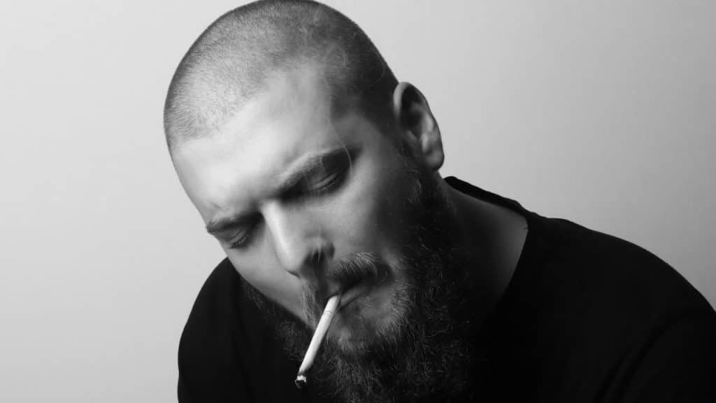 Black and white close-up of a bearded man with eyes closed smoking a lit cigarette.
