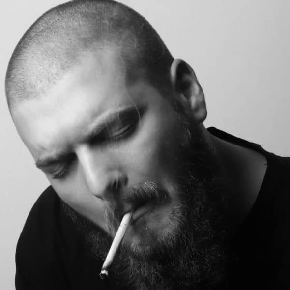 Black and white close-up of a bearded man with eyes closed smoking a lit cigarette.