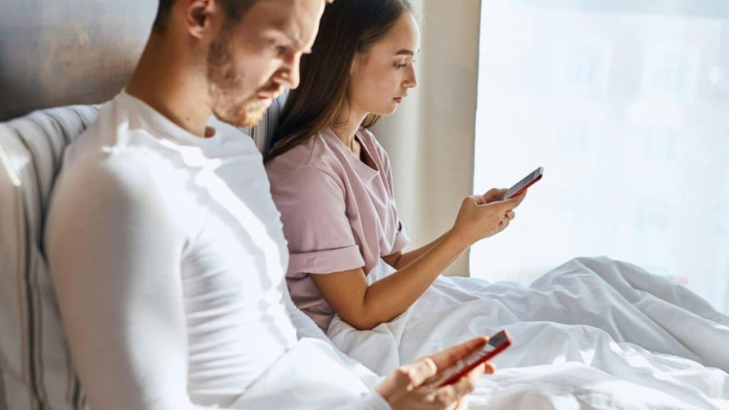 A man and woman sitting in bed together while both look at their smartphones.