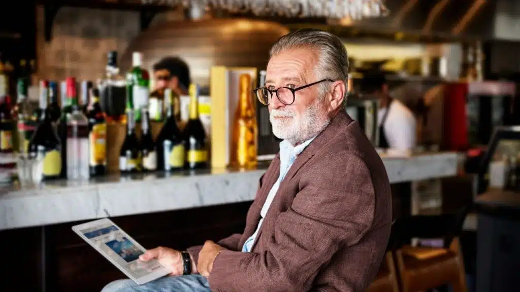 An older man with glasses sitting at a bar holding a tablet and looking away.
