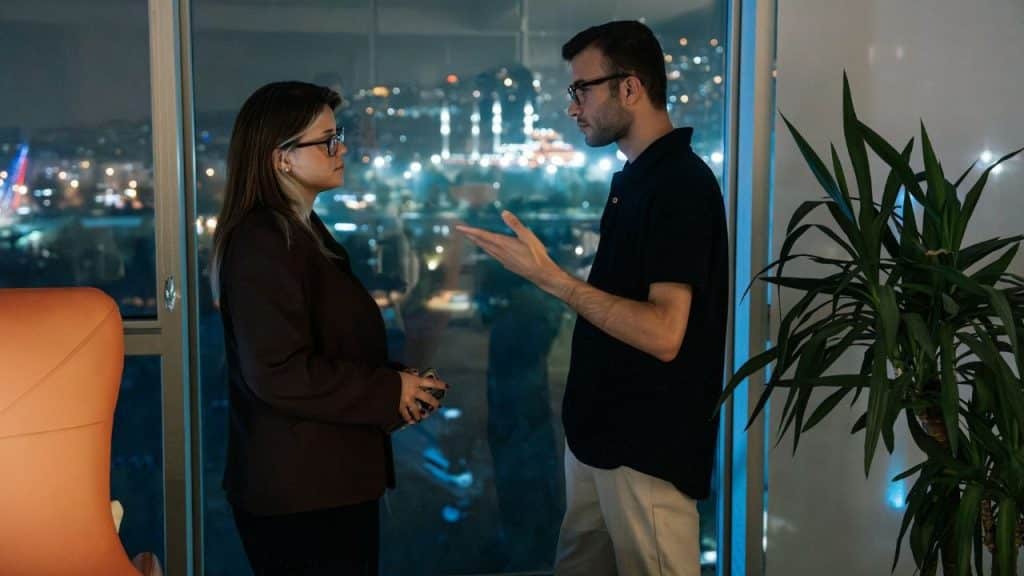 A man and woman standing near a window at night talking and gesturing with hands.