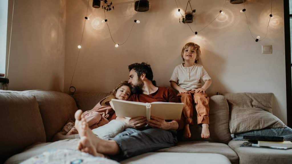 A parent reading a book with two young children on a couch under soft lights.