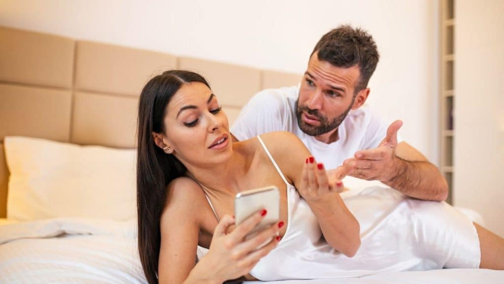 A woman lies on a bed looking at a phone while a man gestures behind her.