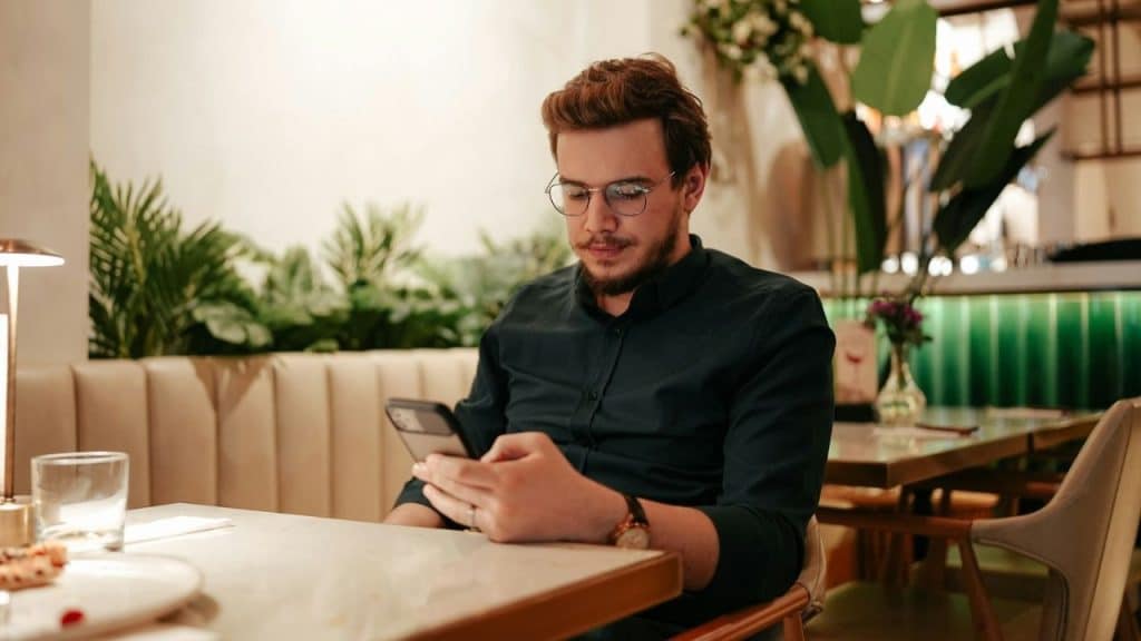 A man wearing glasses sits alone at a restaurant table looking at his mobile phone.