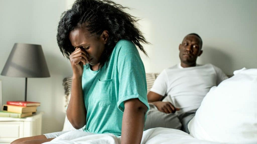 A woman sits on a bed crying into her hand while a man sits behind