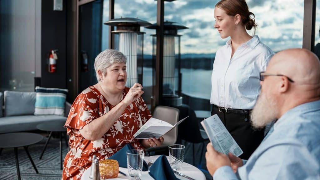 A woman in a floral shirt speaks to a server while holding an open menu.