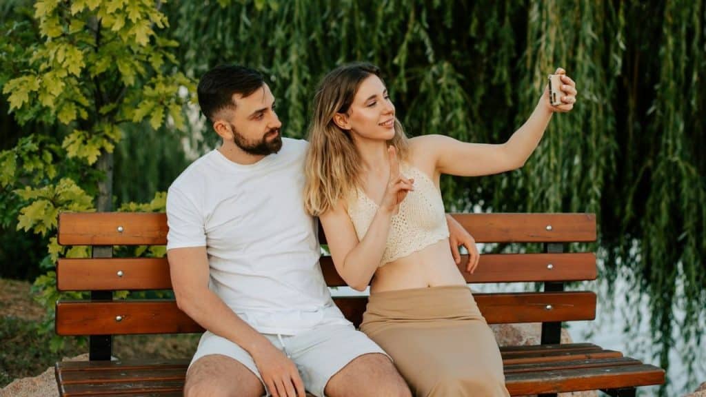 A man and woman sit on a park bench while the woman takes a selfie.