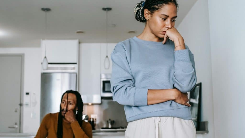A woman in a blue sweatshirt stands with a pensive expression while a man sits behind.