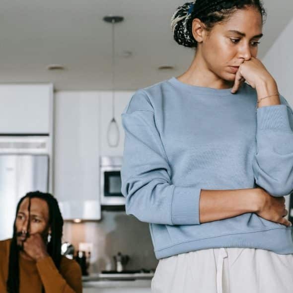 A woman in a blue sweatshirt stands with a pensive expression while a man sits behind.