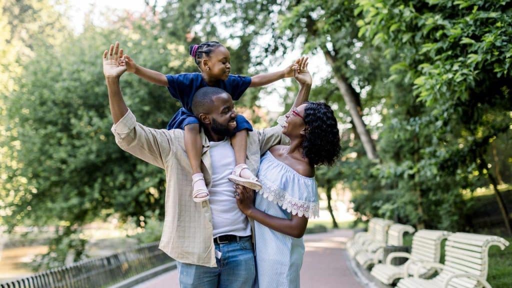 A joyful family walking in a park with a child on an adultโs shoulders.