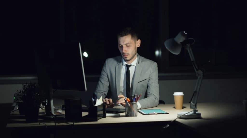 A man in a grey suit and tie works at a computer in a dark room.