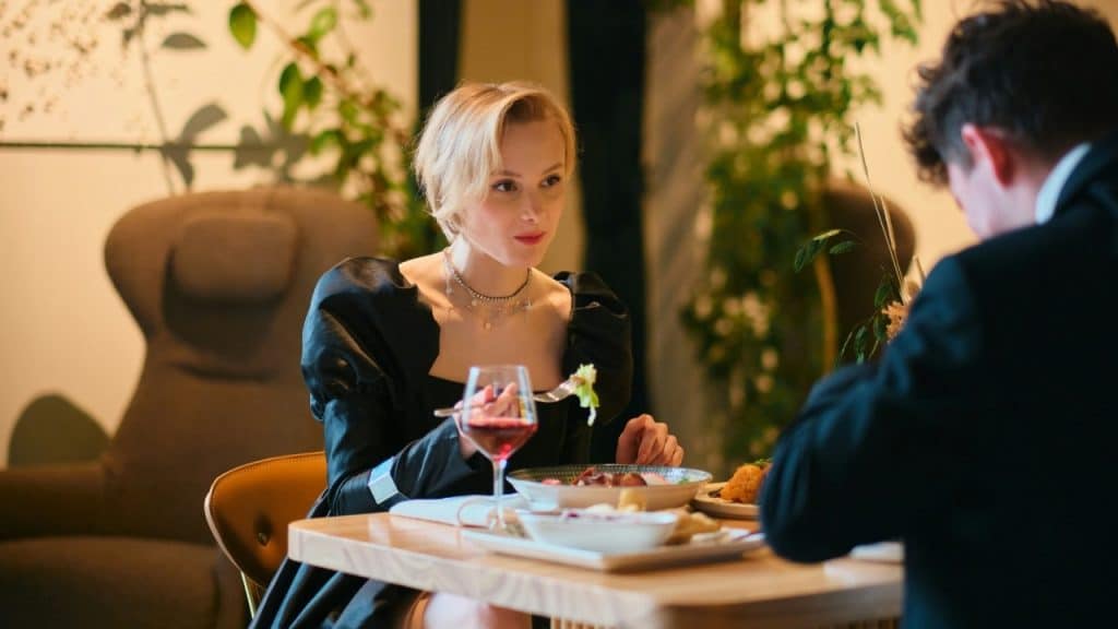 A woman in a black dress looks across a dinner table at a man’s blurred shoulder.