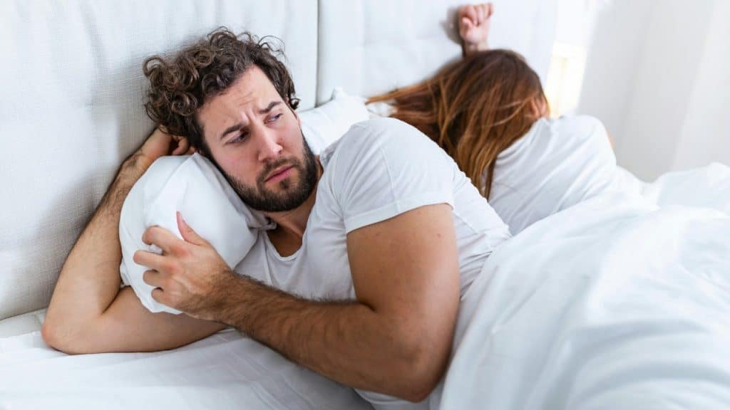 A man with curly hair lies in bed looking away from a woman turned away.