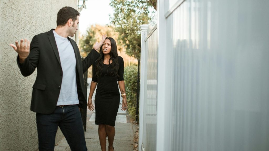 A man and woman stand in a narrow outdoor walkway while gesturing during a conversation.