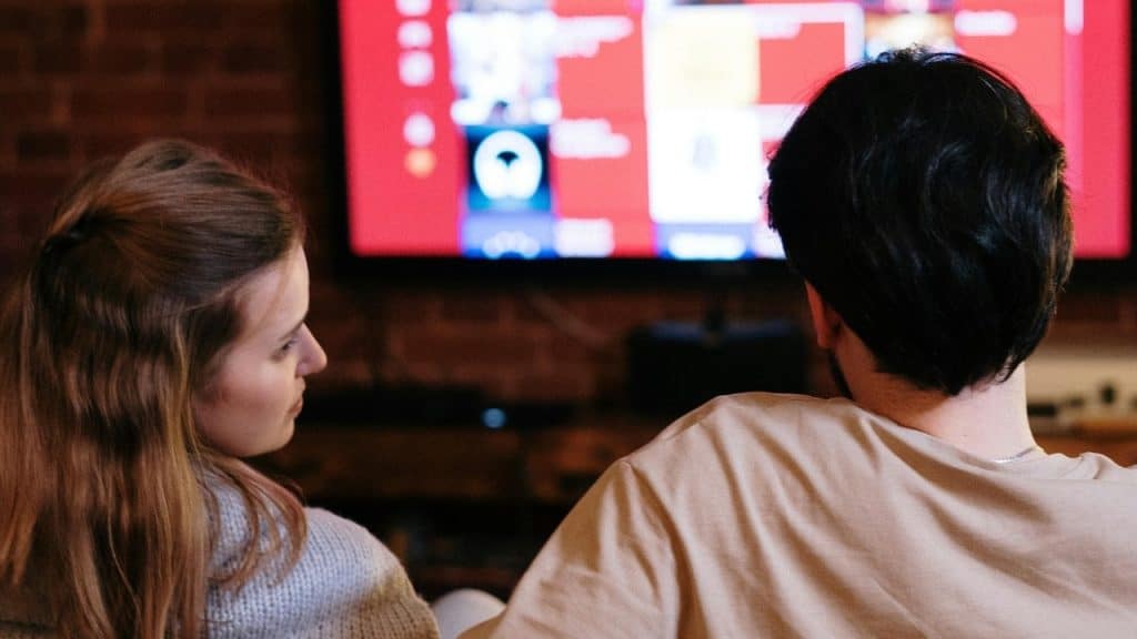A woman looks toward a man’s back while they both face a large, bright television screen.