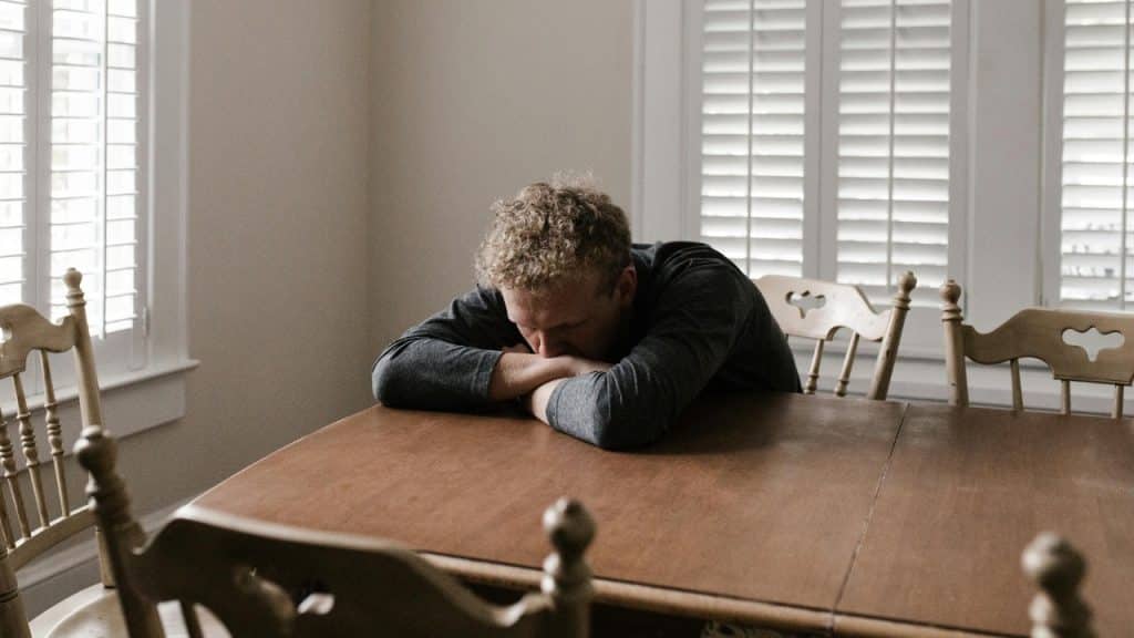 A man with curly hair rests his head on his arms on a wooden dining table.