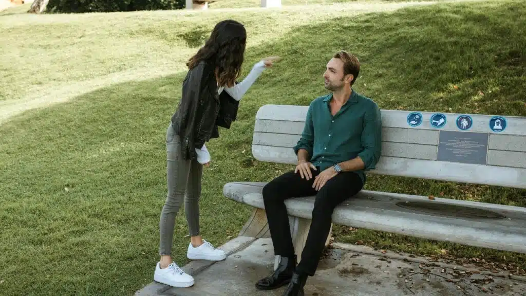 A woman in a leather jacket points toward a man sitting on a public park bench.