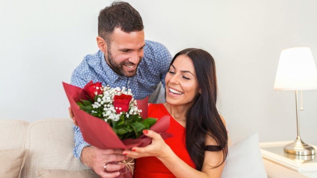 A man presents a bouquet of red roses to a smiling woman sitting on a couch.