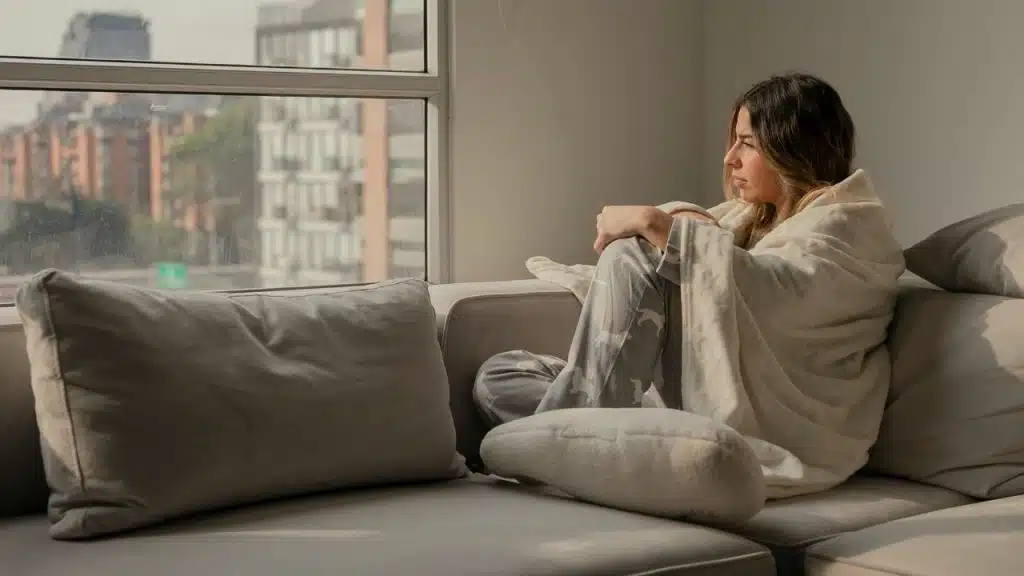 A woman wrapped in a white blanket sits on a grey couch looking out of a window.