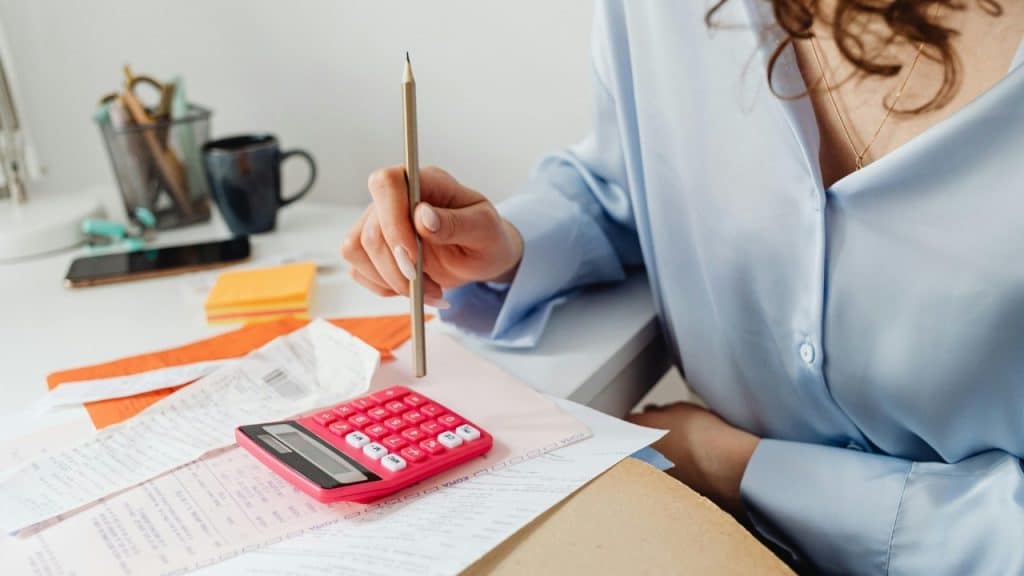 A person uses a pink calculator and a pencil over a pile of white paper receipts.
