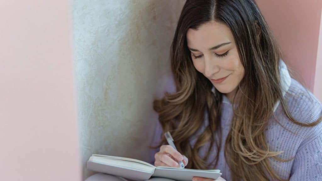 A woman with long hair looks down and writes in a notebook with a grey pen.