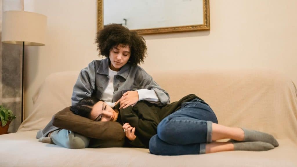 One woman rests her head on the lap of another woman while lying on a couch.