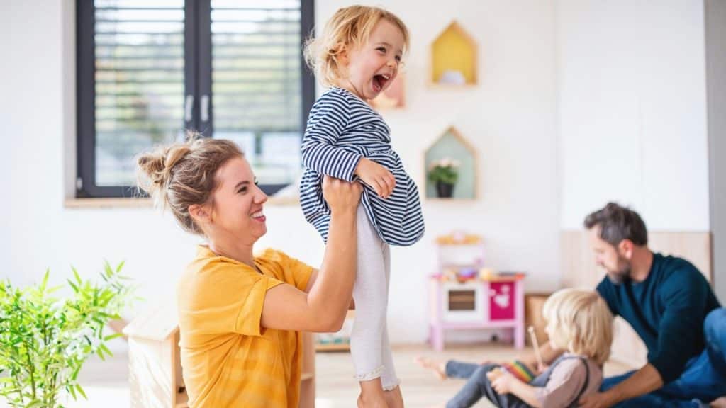 A woman lifts a laughing toddler in a bright room while a man sits nearby playing.