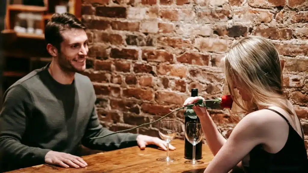 A man and woman sit at a wooden table against a brick wall with a rose.