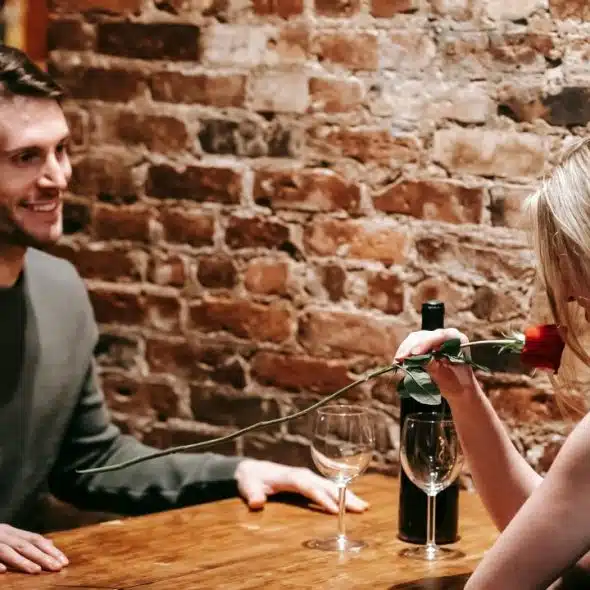 A man and woman sit at a wooden table against a brick wall with a rose.