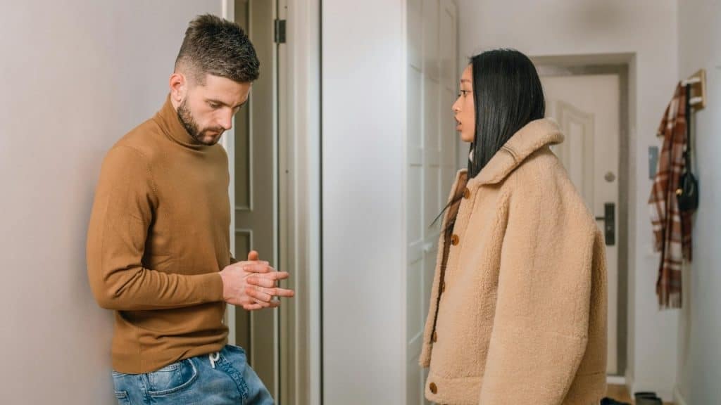 A woman in a tan coat talks to a man in a brown turtleneck in a hallway.