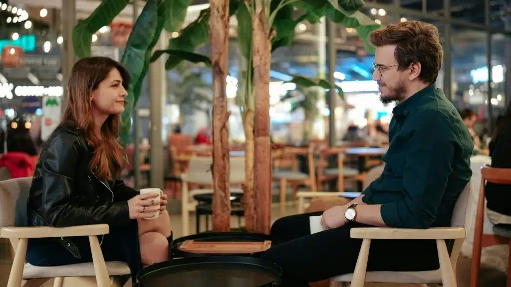 A man and woman sit in chairs at a cafe facing each other with coffee cups.