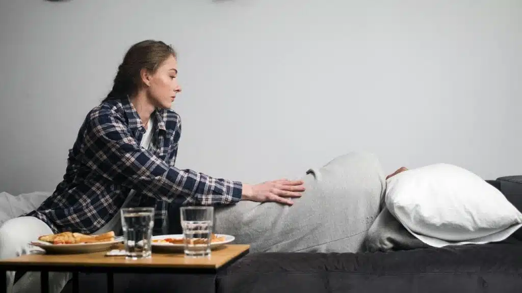 A woman in a plaid shirt sits on a bed touching the back of a person.