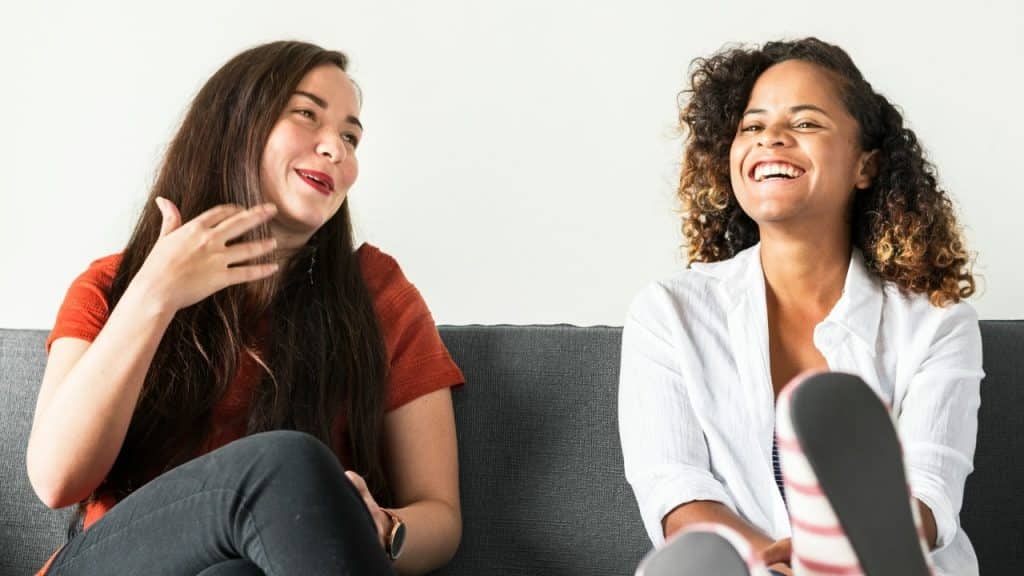 Two women sit on a dark grey couch indoors, laughing and talking with one another.