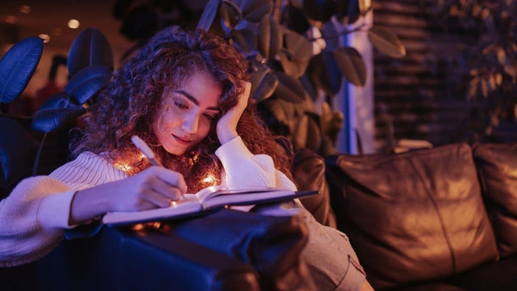 A woman with curly hair writes in a book under blue and orange ambient indoor lighting.