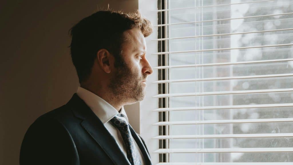 Side profile of a bearded man in a suit looking out through horizontal window blinds.