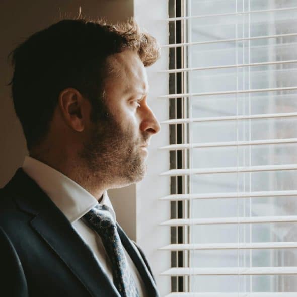 Side profile of a bearded man in a suit looking out through horizontal window blinds.
