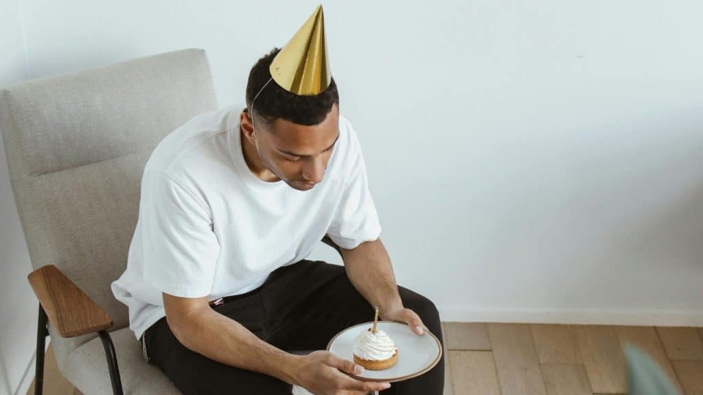 A man wearing a gold party hat sits alone holding a small cake with a candle