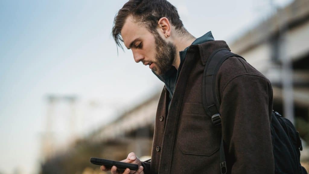 A man with a backpack and brown jacket looks down at a smartphone held in his hand.